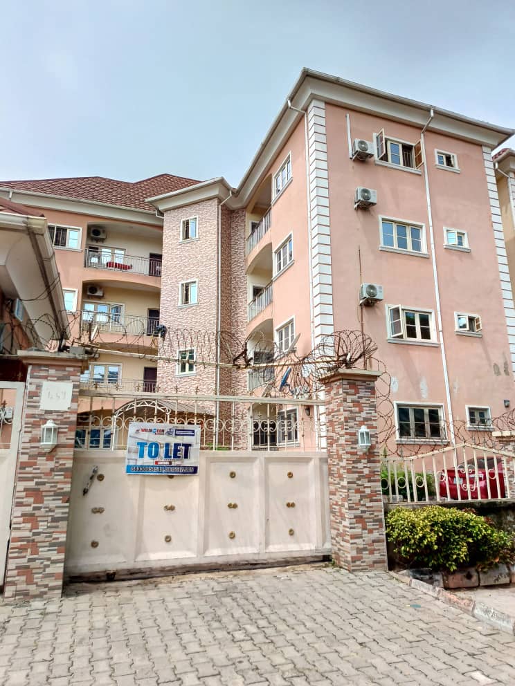 modern commercial district in Abuja, Nigeria, featuring a variety of office buildings, retail shops, and industrial warehouses. The architecture is contemporary and well-maintained, reflecting a thriving business environment. Sunlight casts sharp shadows, highlighting the textures of concrete, glass, and metal. The scene includes well-paved roads, some pedestrian activity, and a clear blue sky.