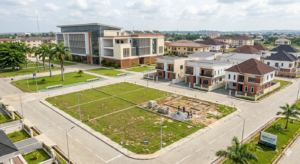 An aerial view photograph capturing the developing Walexdot Real Estate estate in Nigeria on a bright, sunny day. The master-planned gated community features distinct zones: a modern university campus building on the left, an established phase of completed modern houses in the middle ground, and a new 'Walexdot' phase in the foreground. This new phase showcases several identical, brown-roofed prototype houses and dozens of cleared, marked-out plots ready for development. Wide, freshly paved asphalt roads with white lane markings and sidewalks connect the phases. Palm trees and landscaped green areas are interspersed throughout the development. In the foreground near the entrance, prominent signage is visible, including a main stone-based monument sign with the 'Walexdot' brand and smaller directional 'Phase 1' signs. The background shows a sprawling, mature residential town under a partly cloudy sky.