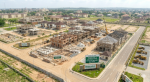 Buying Off-Plan Property in Nigeria, An aerial perspective photograph of a bustling, active construction site within the master-planned Walexdot estate on a clear day. In the center, multiple modern houses are visibly underway in various stages of completion, showcasing exposed brickwork, concrete foundations, and wooden roof trusses. Active construction machinery, including yellow excavators, a crane, and concrete rollers, are operating throughout the grounds. Near the freshly paved asphalt road entrance stands a prominent stone monument sign bearing the "Walexdot" logo, right next to a large billboard that reads "NOW SELLING: PHASE 2 & 3 (OFF-PLAN UNITS); ESTIMATED DELIVERY: Q3 2025." Completed brown-roofed houses stand further back, contrasting with the active build zone.